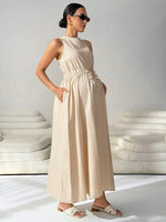Load image into Gallery viewer, A woman modeling a sleeveless, high-neck maxi dress in a light beige or cream color. The dress features a gathered elastic waist with a matching fabric tie belt and side pockets.
