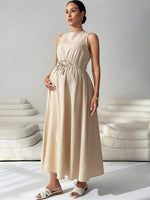 Load image into Gallery viewer, A pregnant woman wearing a sleeveless, light beige maxi dress with a gathered waist and tie detail, standing in a minimalist studio setting with white modern sofas in the background.
