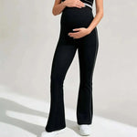 Load image into Gallery viewer, A pregnant model wearing black maternity flare pants with white side stripes and a matching black top, standing against a light gray studio background.
