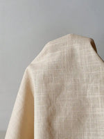Load image into Gallery viewer, A close-up of draped fabric with a fine, subtle grid or crosshatch texture in a warm beige tone against a plain light gray background.
