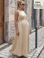 Load image into Gallery viewer, A pregnant woman wearing a sleeveless, floor-length beige maternity dress with a tie waist, standing on a cobblestone street next to a stone wall. A text overlay in the top right corner reads '100% Cotton Comfortable'.
